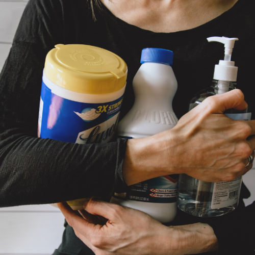 woman wearing a black long-sleeved shirt holding Lysol wipes, bleach, and hand sanitizer