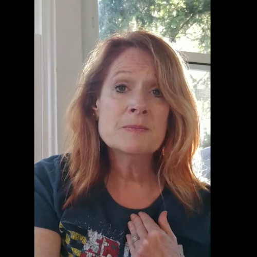 Tracey Tiernan sitting in front of a window wearing a blue shirt and looking into the camera with her left hand on her heart