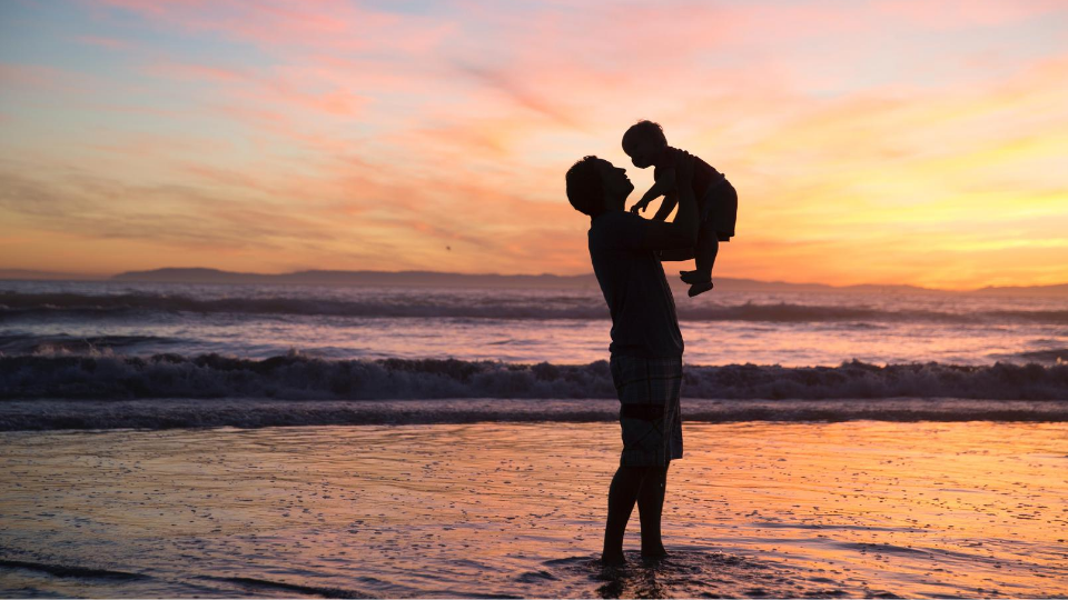 Shadow of dad holding a little boy up in the air in front of an ocean and orange and yellow sunset