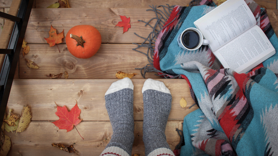 person's feet pictured on light brown steps wearing grey socks with a pumpkin to the left and a book and blanket with coffee to the right and red and orange leaves scattered around the feet