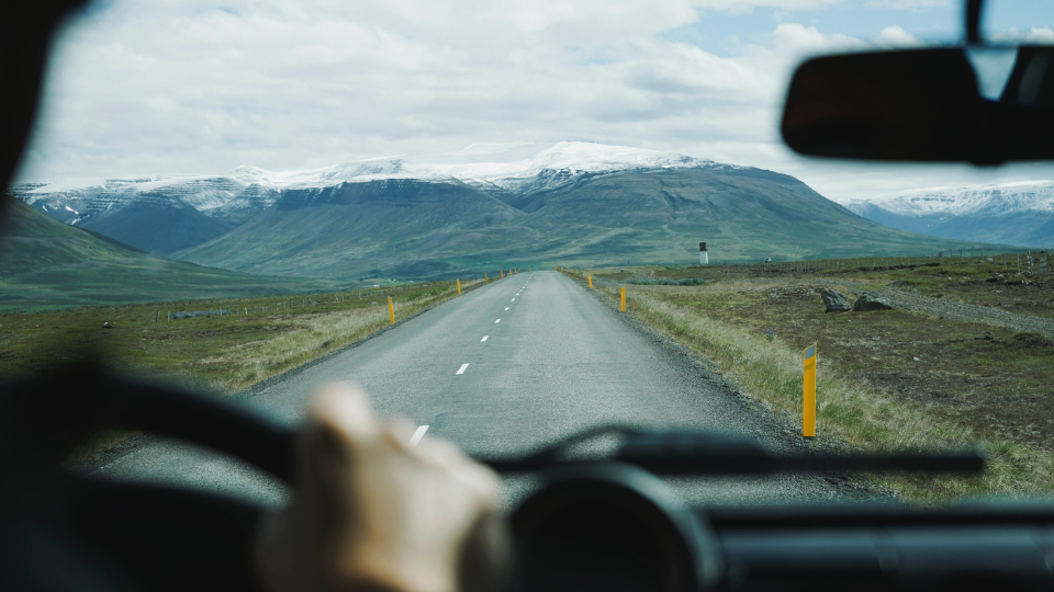 hands resting on a steering wheel while driving down a long paved road into snowcapped mountains