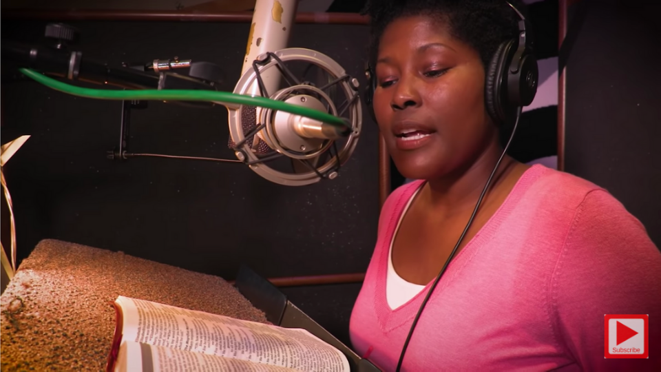 Black woman wearing a pink sweater standing behind a microphone with headphones on as she looks down at a Bible