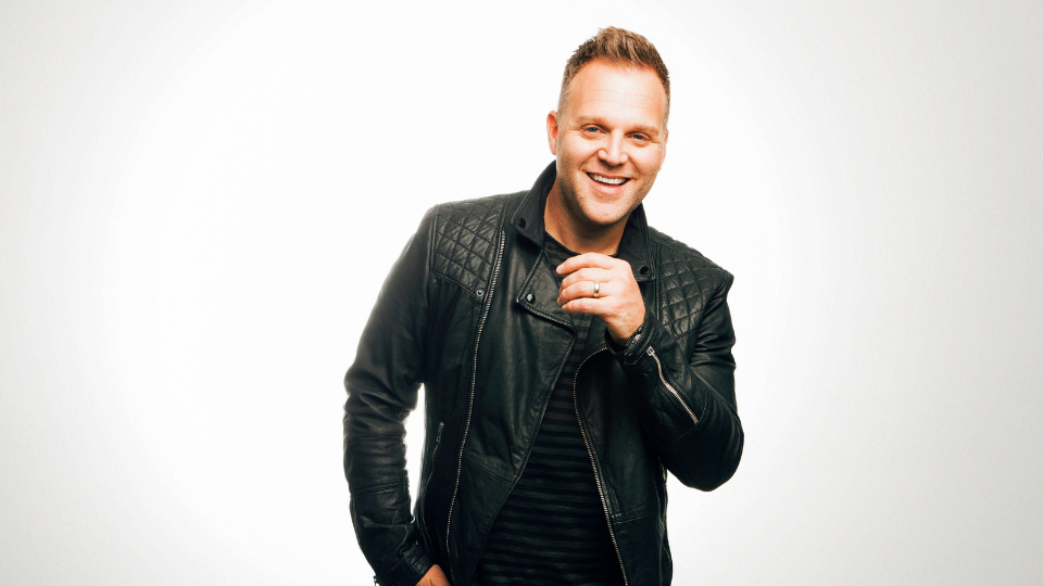 Matthew West smiling while lifting his left hand up towards his chin and wearing a black leather jacket He is standing in front of white backdrop