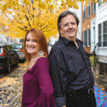 Tracey and Mike standing back to back on a side street with a yellow tree in the background and yellow leaves scattered on the ground