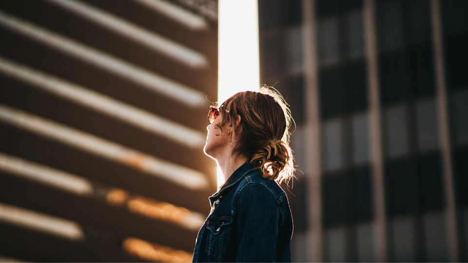 woman with low bun and glasses looking up at sun peaking between two city buildings