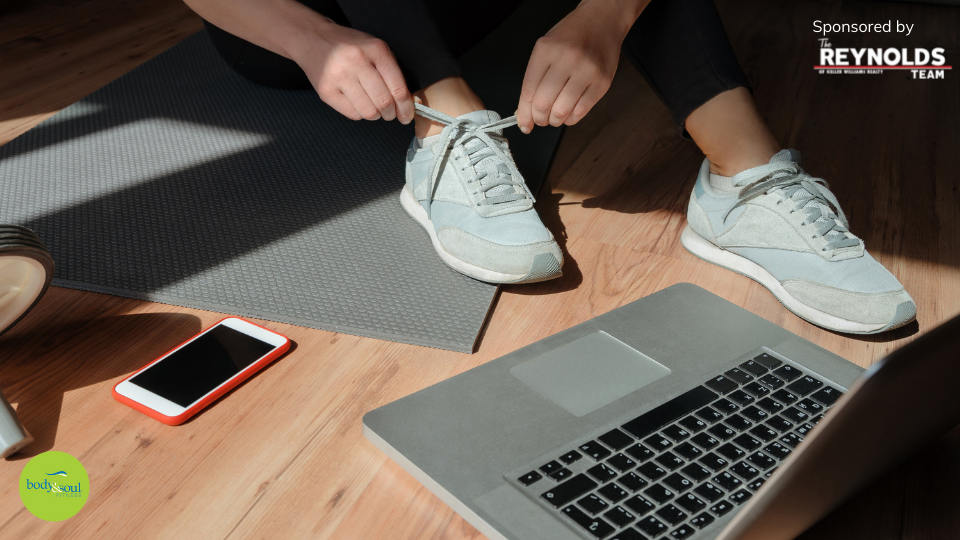 person tying their grey tennis shoes while sitting on a grey yoga mat in front of a laptop on the floor