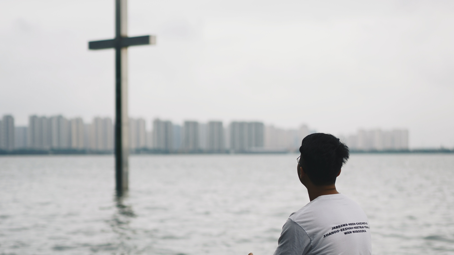 man with black hair with his back to the camera as he looks out at a cross placed in a river with a city skyline in the background