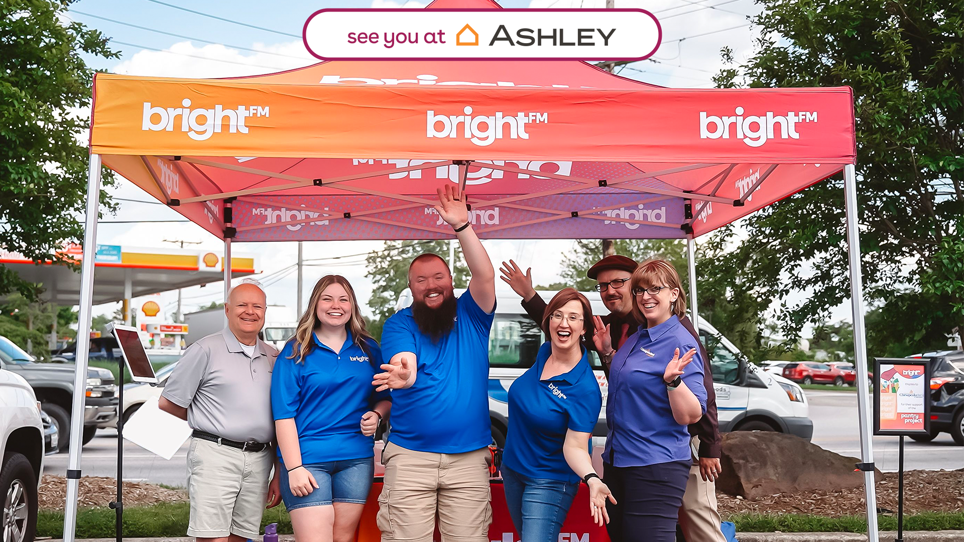 BRIGHT-FM team members smiling and lifting their hands up while standing under a pink and orange BRIGHT-FM tent