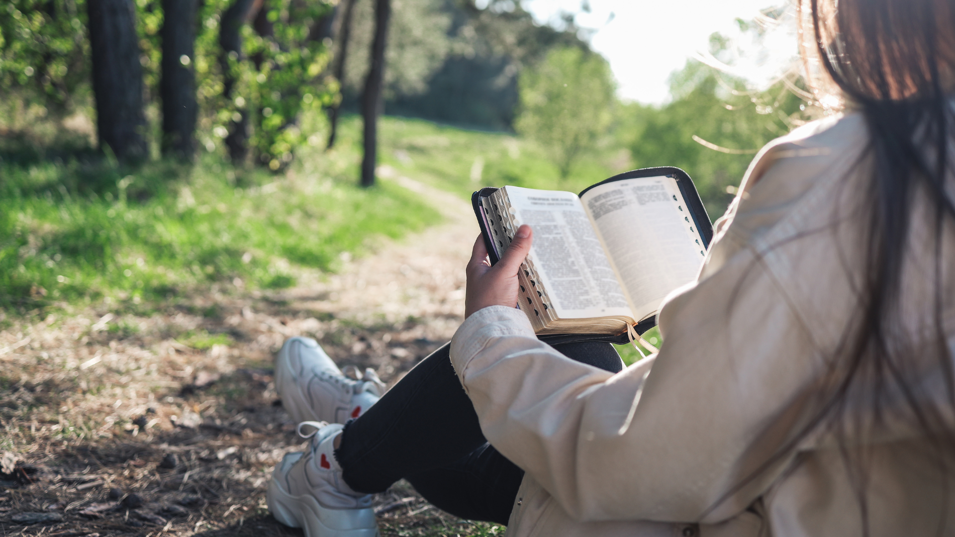 woman sitting against a tree on a path through a forest while she reads her Bible