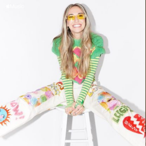Lauren Daigle smiling and kicking her legs out as she sits on a white stool while wearing white pants with colorful drawings and a bright green shirt. The entire outfit is topped off transparent yellow glasses