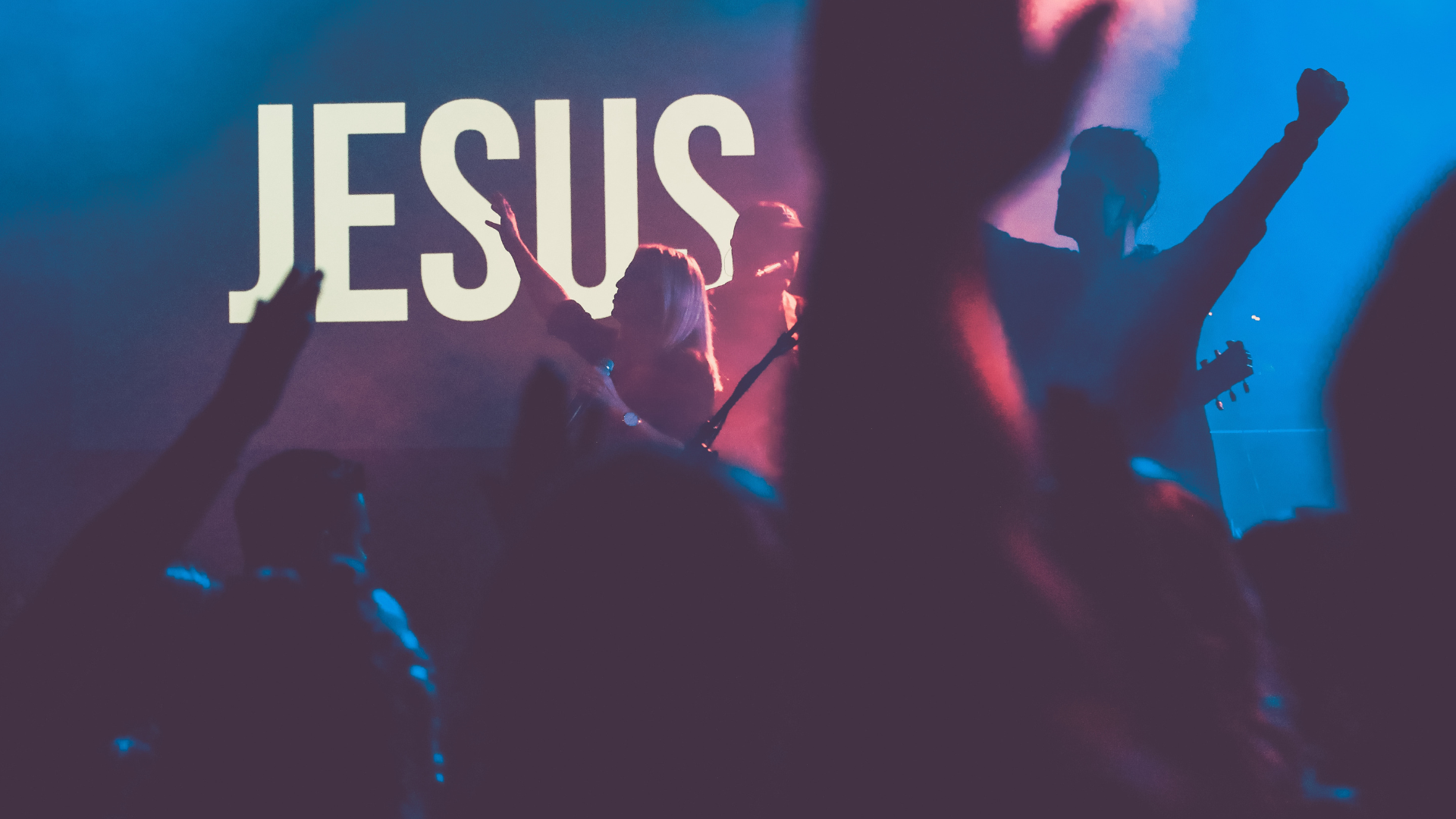 People standing at a worship event with their hands raised up in praise and the name of "Jesus" displayed on the screen on stage