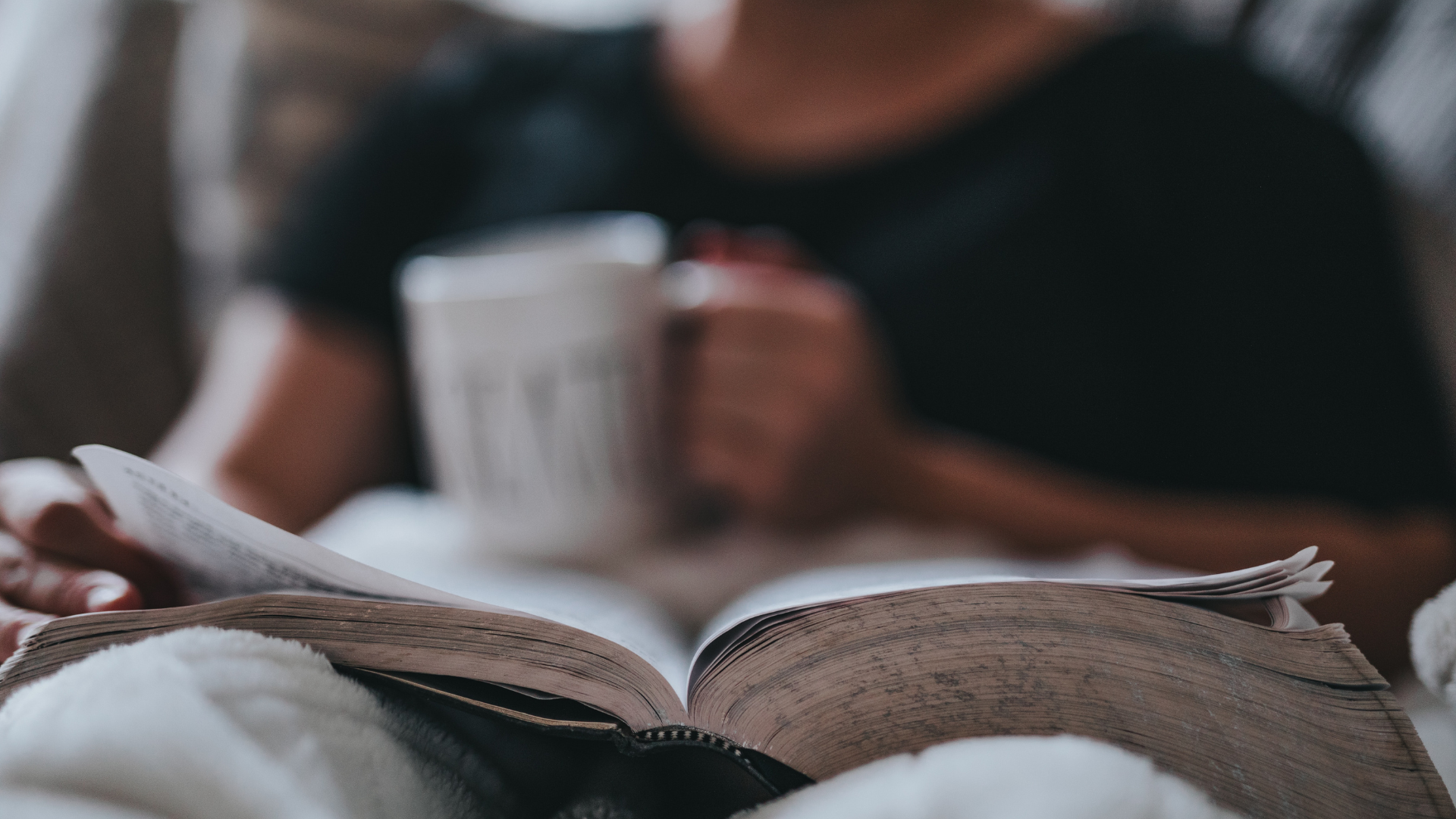 Person sitting in a chair reading the Bible while holding a mug and snuggling in a blanket